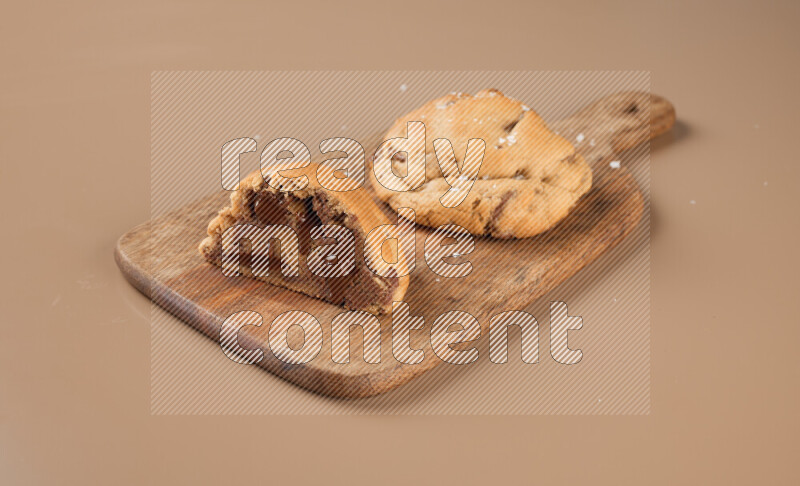 a chocolate chip cookie with another one cut in half on a wooden cutting board on a brown background