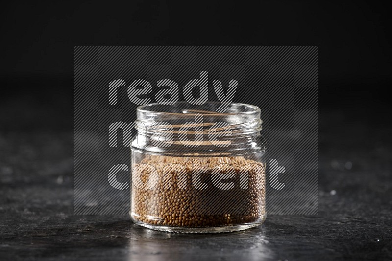 A glass jar full of mustard seeds on a textured black flooring in different angles
