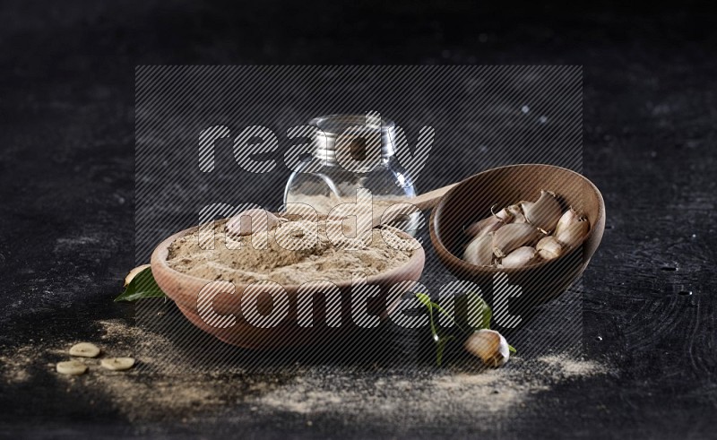 A wooden bowl, spoon and glass spice jar full of garlic powder and a wooden bowl full of garlic cloves on a textured black flooring in different angles