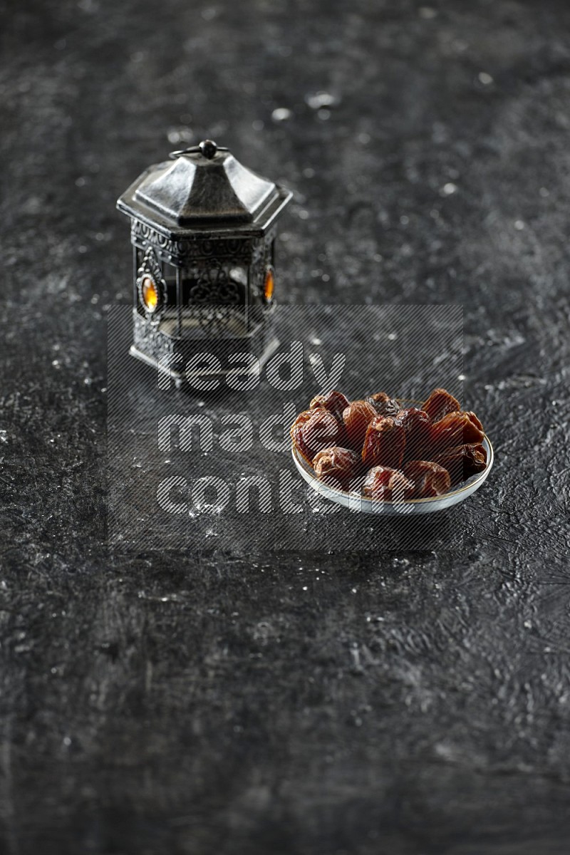 A silver lantern with different drinks, dates, nuts, prayer beads and quran on textured black background