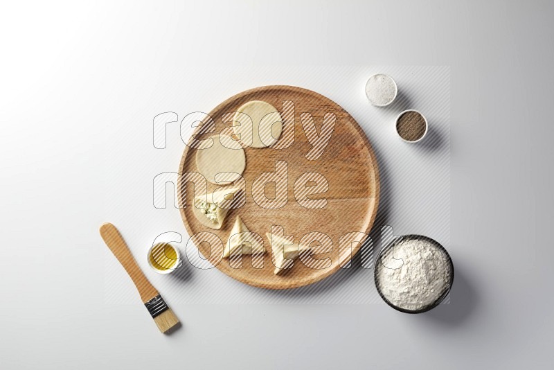 two closed sambosas and one open sambosa filled with cheese while flour, salt, black pepper and oil with oil brush aside in a wooden dish on a white background