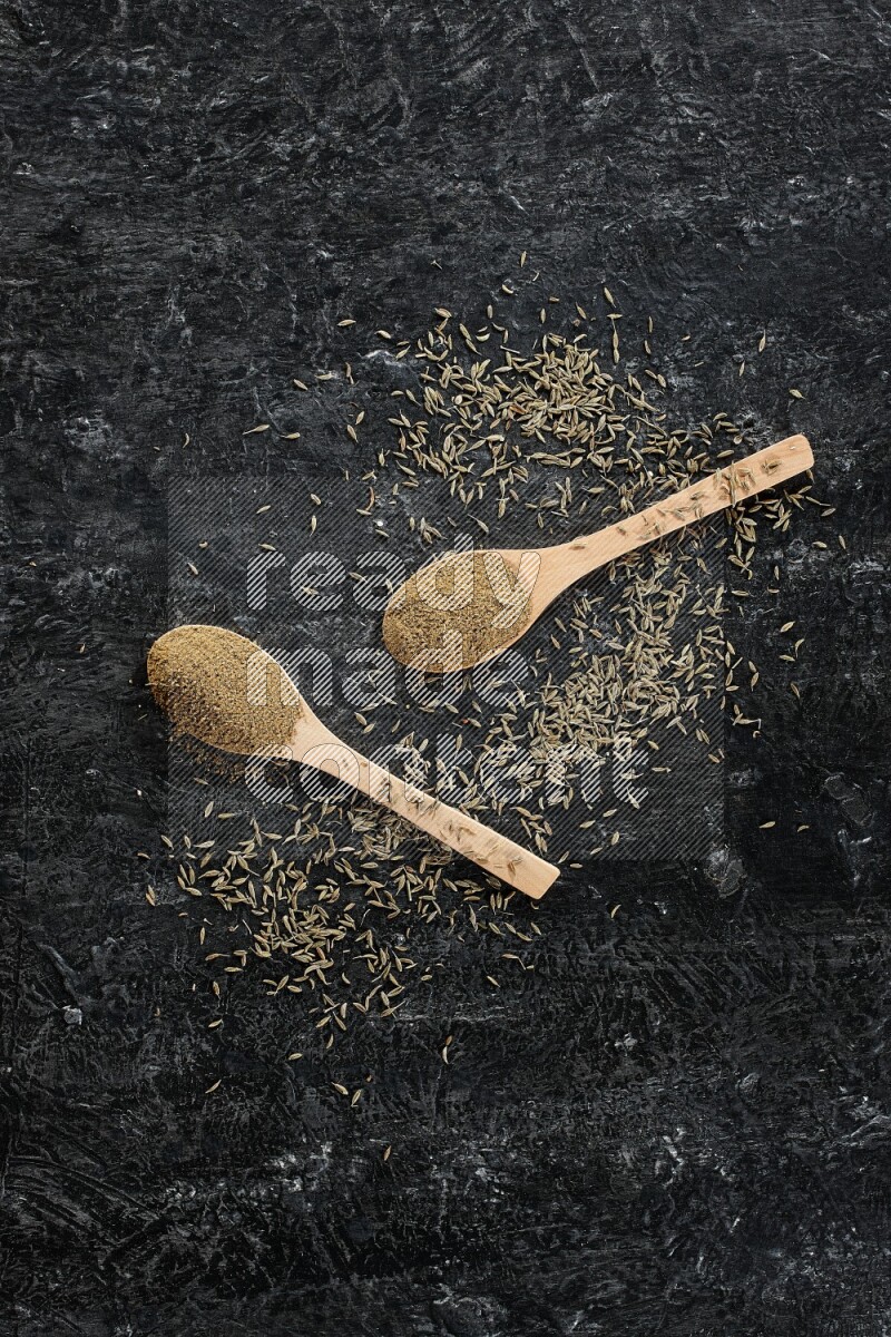 2 wooden spoons full of cumin powder with spreaded seeds on textured black flooring
