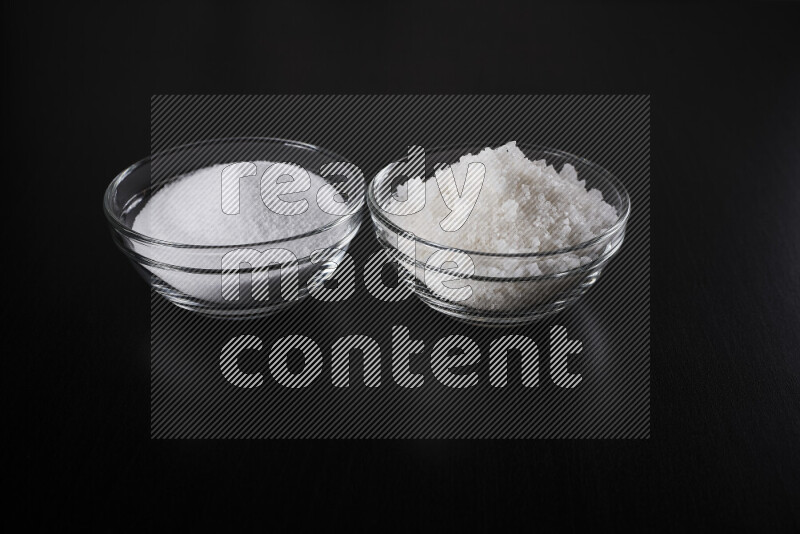 2 glass bowls one is filled with table salt and the other with coarse sea salt on black background