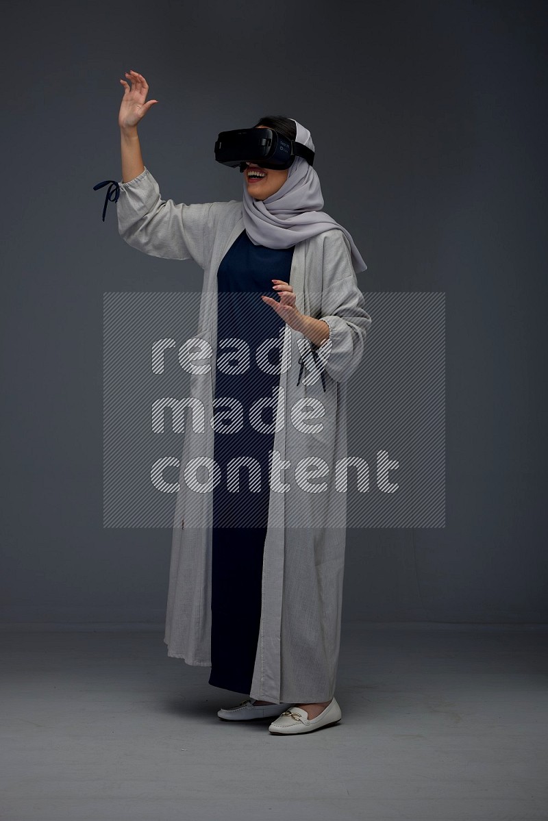 A Saudi woman wearing a light gray Abaya and head scarf standing and wearing VR eye level on a grey background