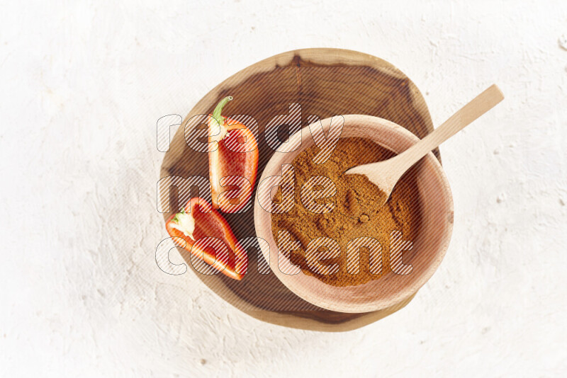 A wooden bowl full of ground paprika powder and sliced red bell pepper beside it all on a wooden tray on white background