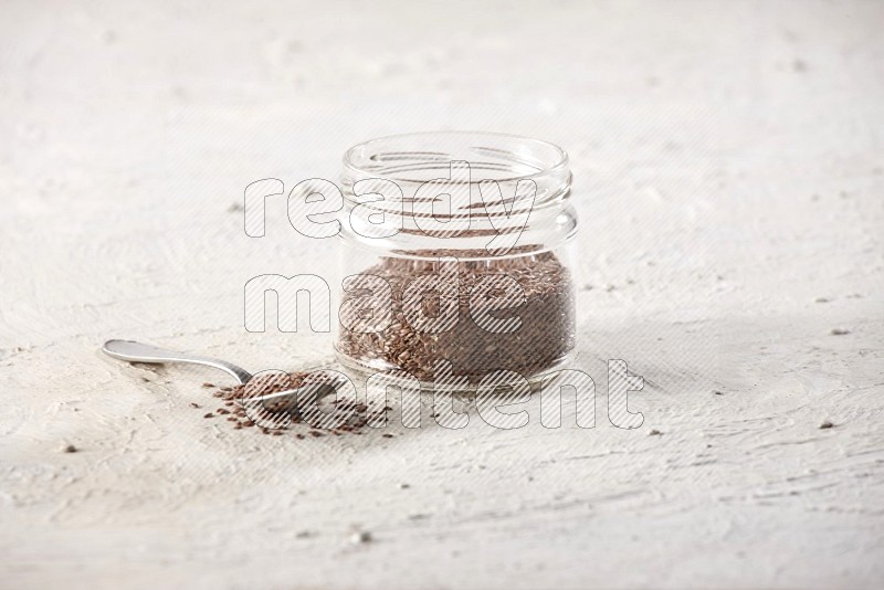A glass jar full of flax seeds with a metal spoon full of the seeds on a textured white flooring