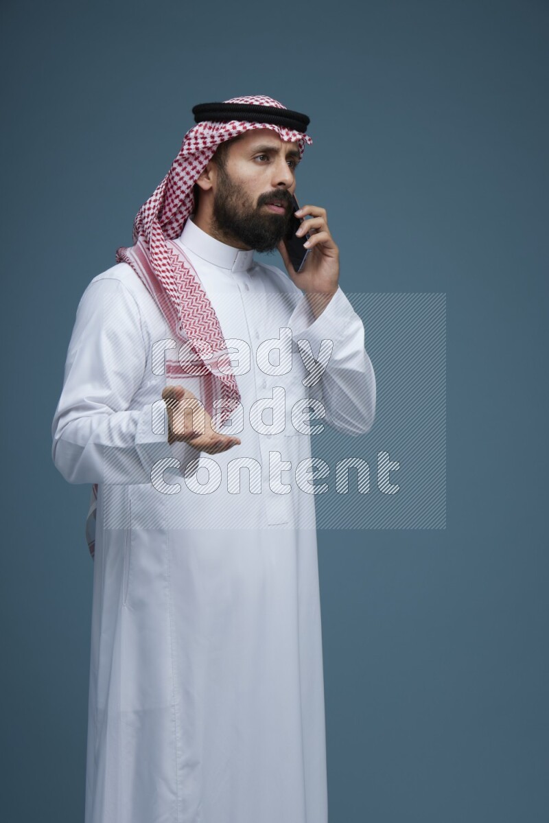 A man having a Call in a blue background wearing Saudi Thob with Shomag
