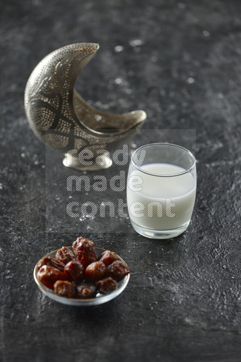 A silver lantern with different drinks, dates, nuts, prayer beads and quran on textured black background