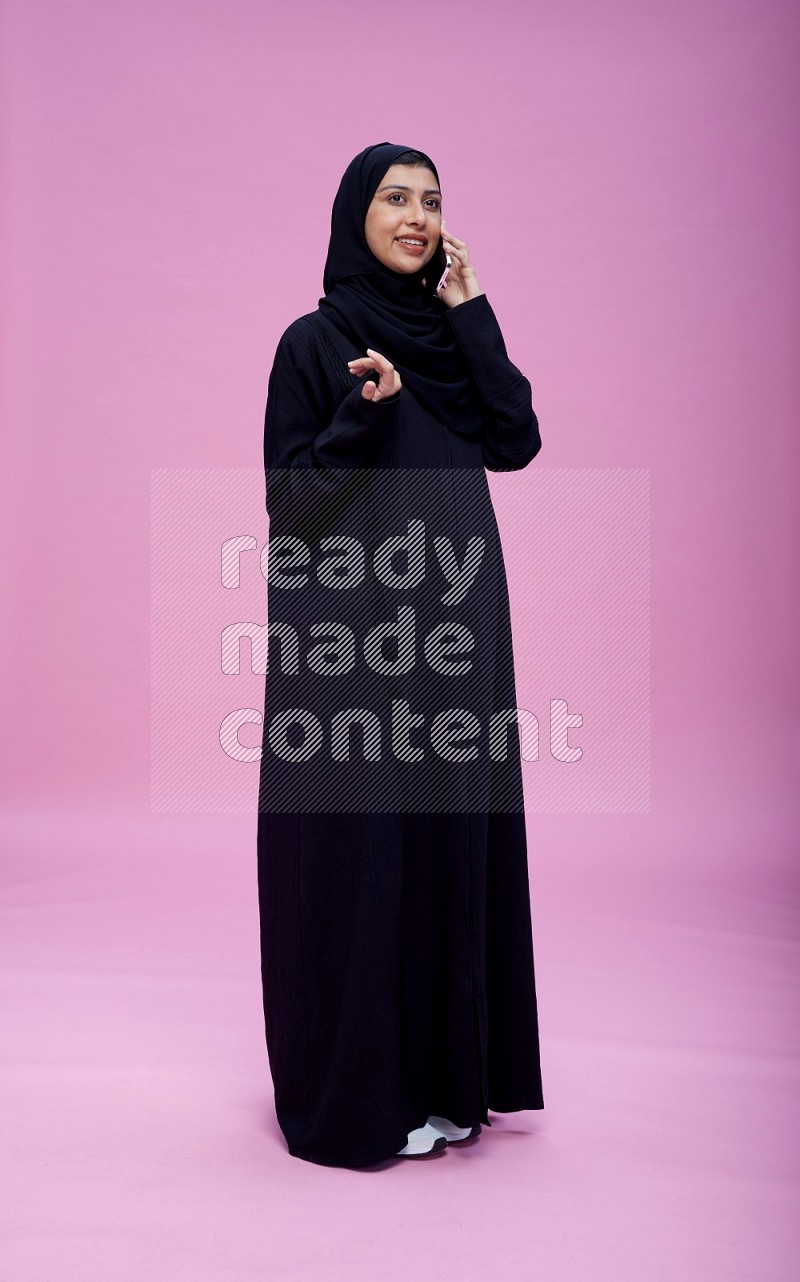 Saudi woman wearing Abaya standing talking on phone on pink background