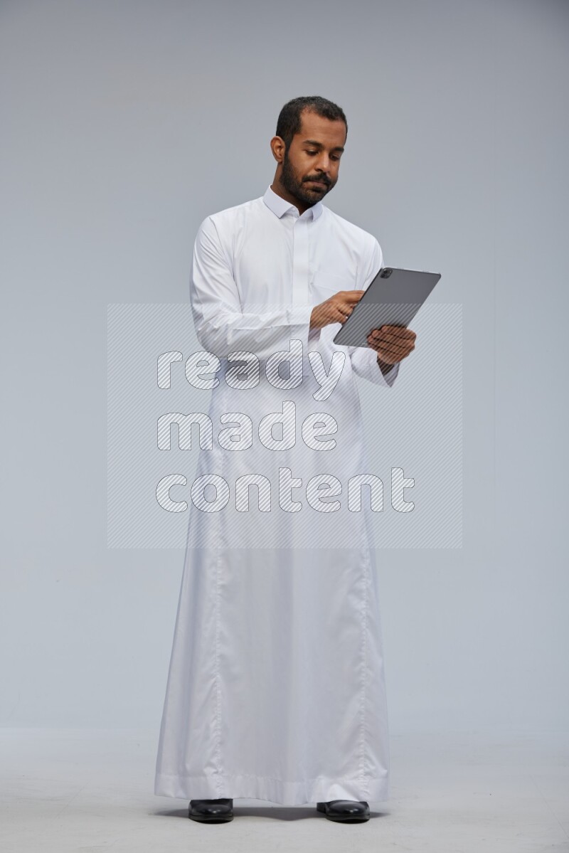 Saudi man wearing Thob standing working on tablet on Gray background
