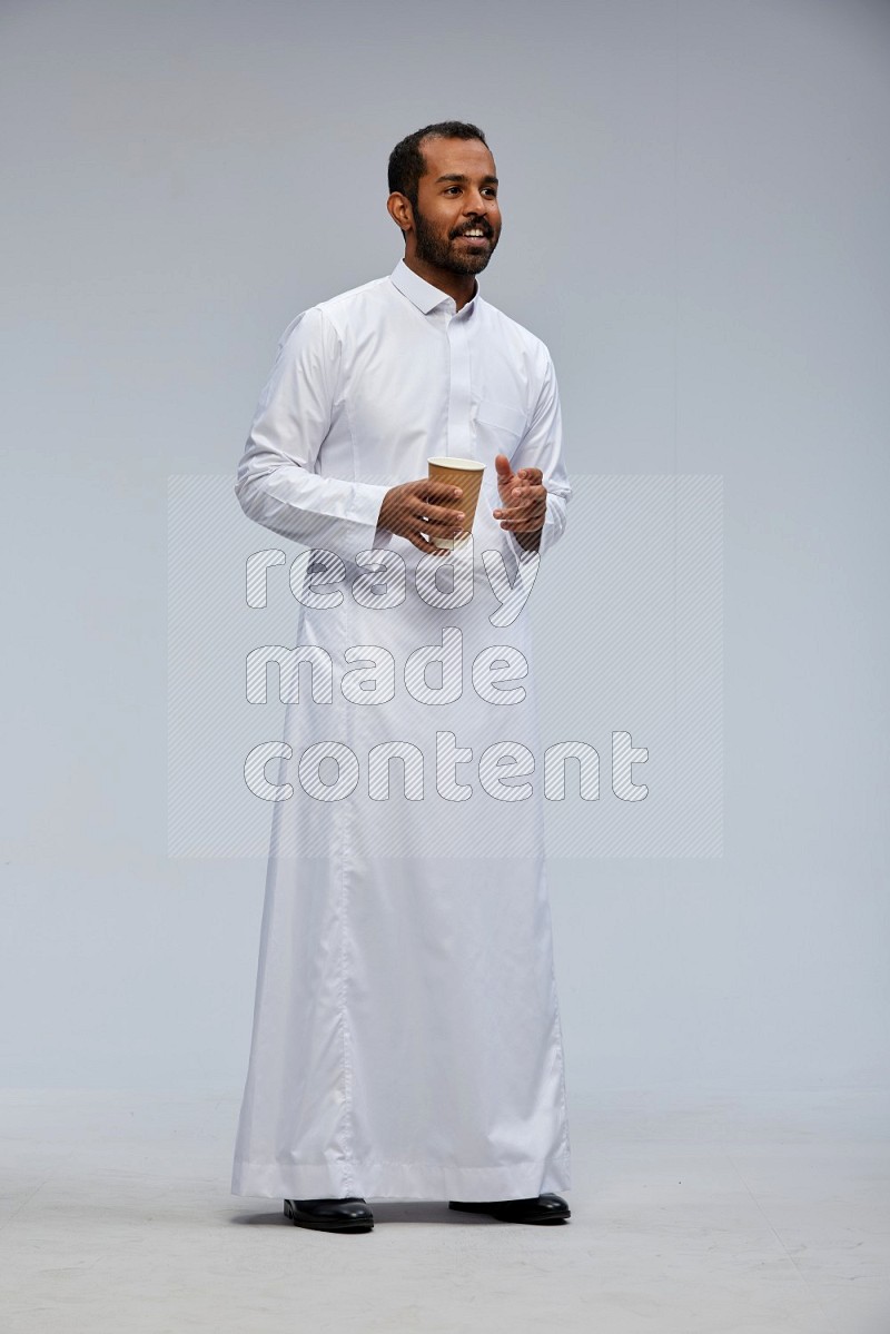 Saudi man wearing thob standing holding paper cup on gray background