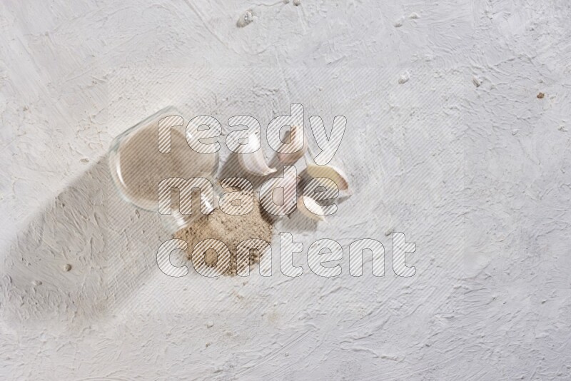 A glass spice jar full of garlic powder flipped over with the powder came out on a textured white flooring