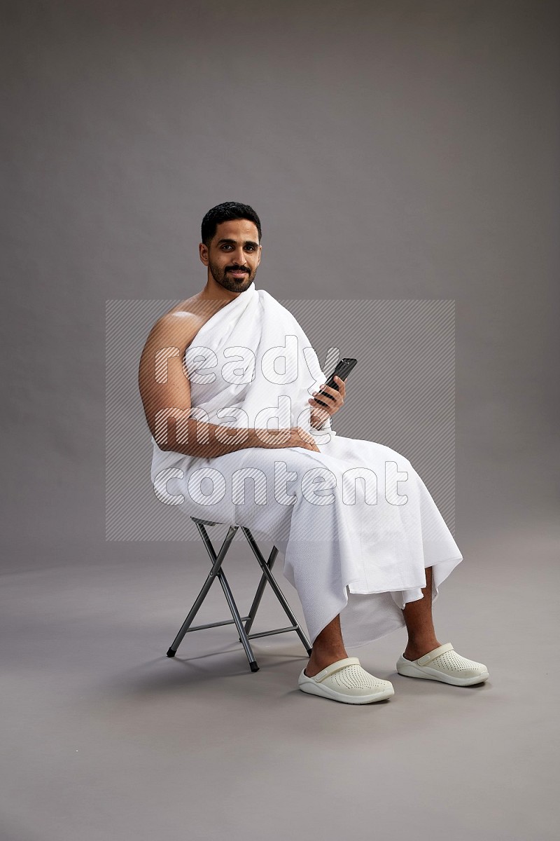 A man wearing Ehram sitting on chair texting on phone on gray background