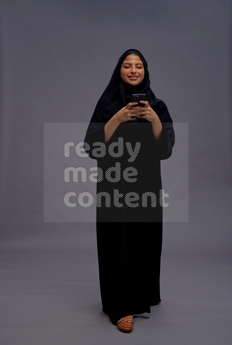 Saudi woman wearing Abaya standing texting on phone on gray background