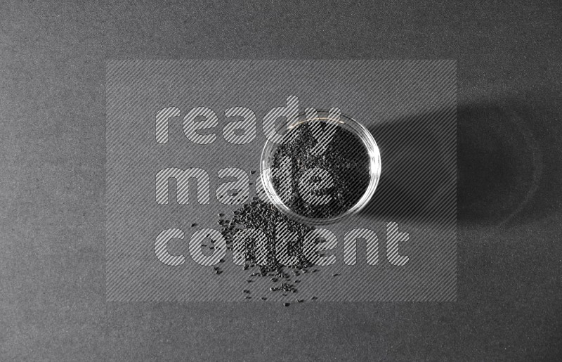 A glass bowl full of black seeds and seeds spreaded beside it on a black flooring