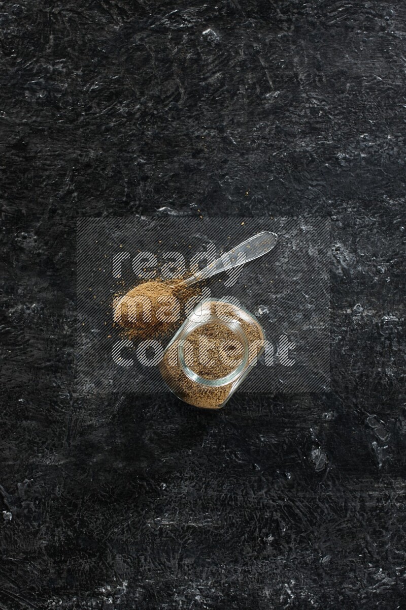 A glass spice jar and a metal spoon full of cumin powder on a textured black flooring