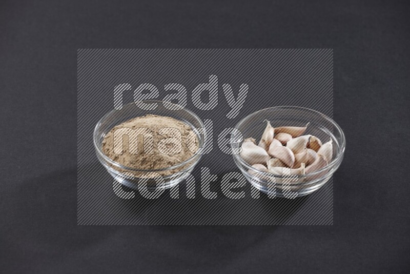 2 glass bowls, one full of garlic powder and the other full of garlic cloves on a black flooring
