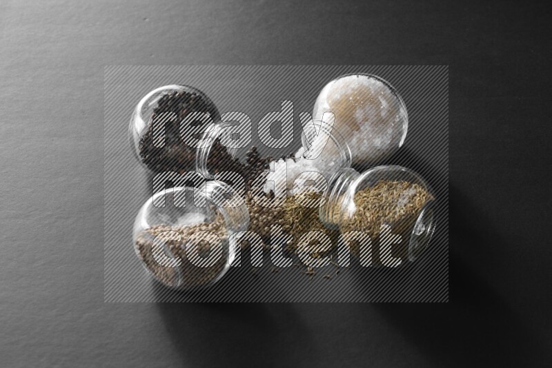 4 glass spice jars full of salt, black peppers, white peppers and cumin on black flooring