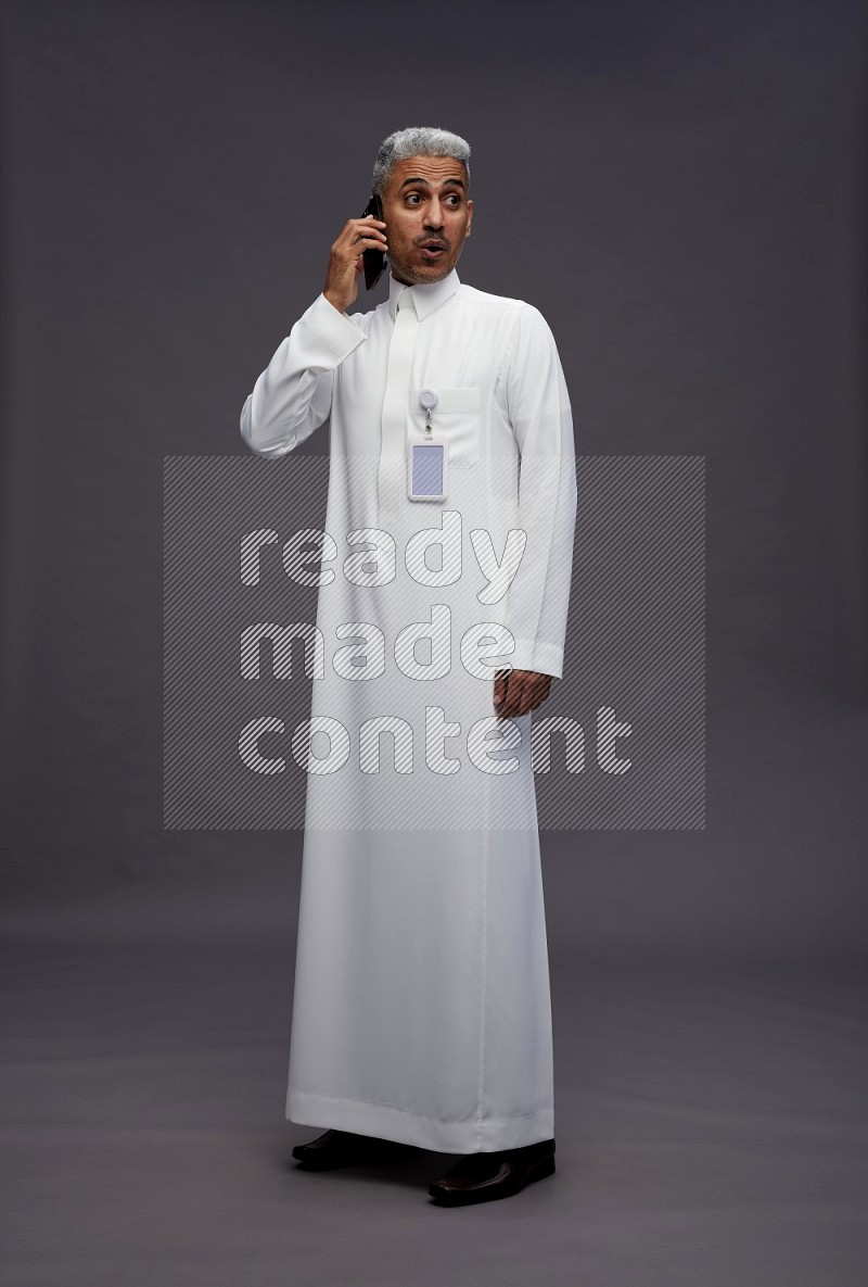 Saudi man wearing thob with pocket employee badge standing talking on phone on gray background