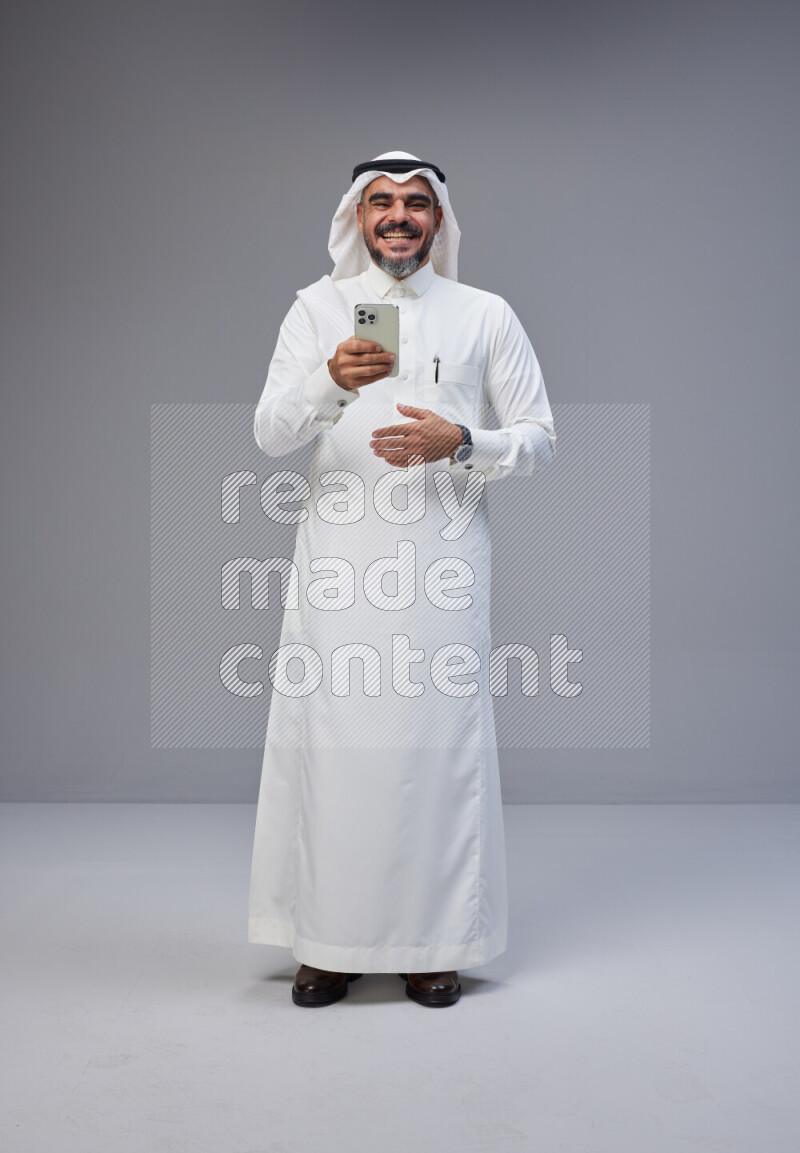 Saudi man Wearing Thob and white Shomag standing texting on phone on Gray background