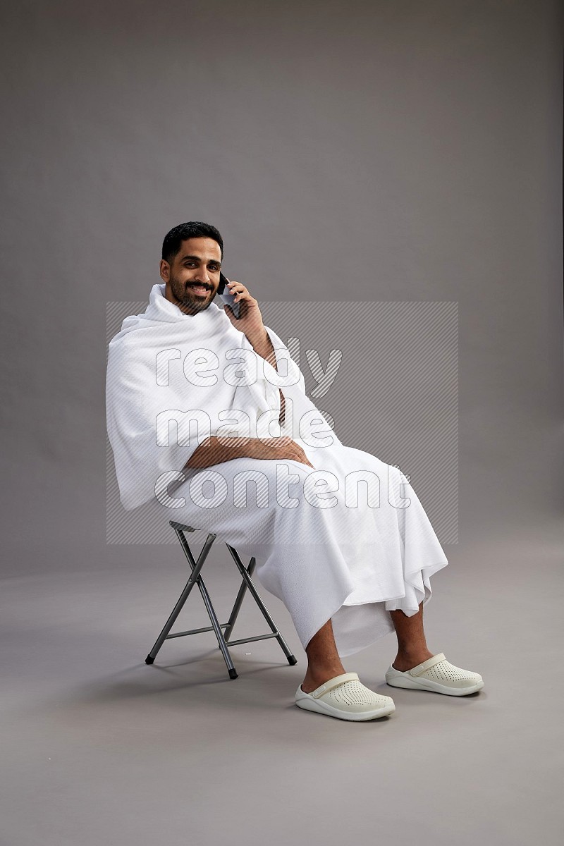 A man wearing Ehram sitting on chair talking on the phone on gray background
