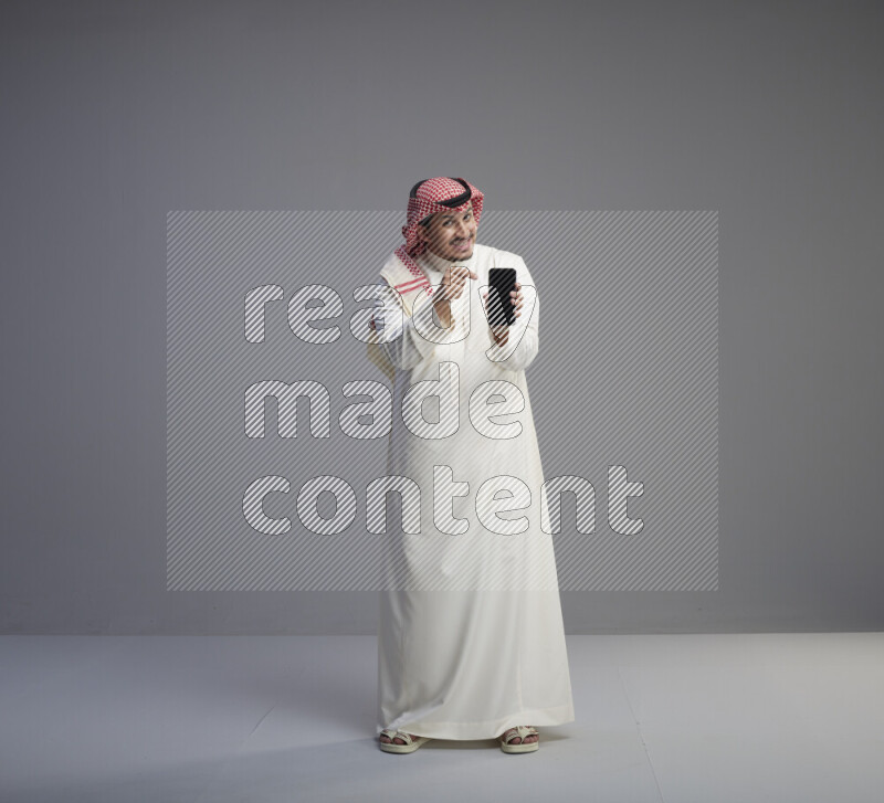 A Saudi man standing wearing thob and red shomag showing phone to camera on gray background