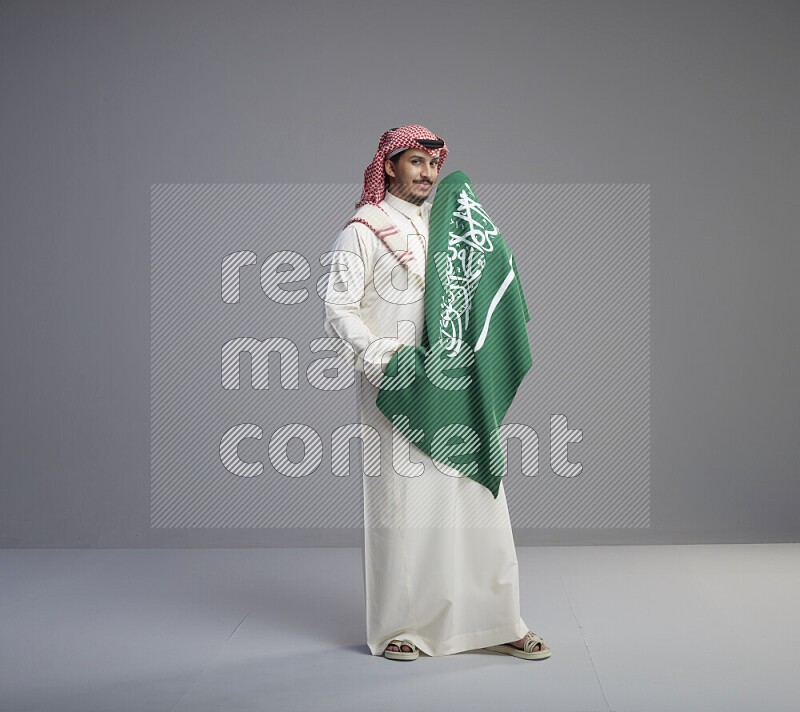 A saudi man standing wearing thob and red shomag holding big saudi flag on gray background