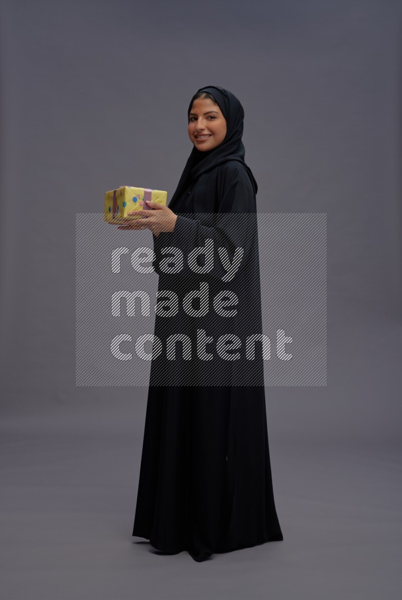 Saudi woman wearing Abaya standing holding gift box on gray background