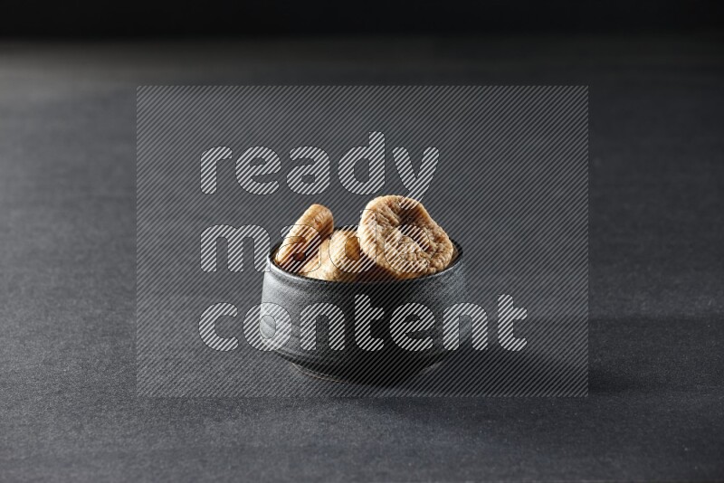 A black pottery bowl full of dried figs on a black background in different angles