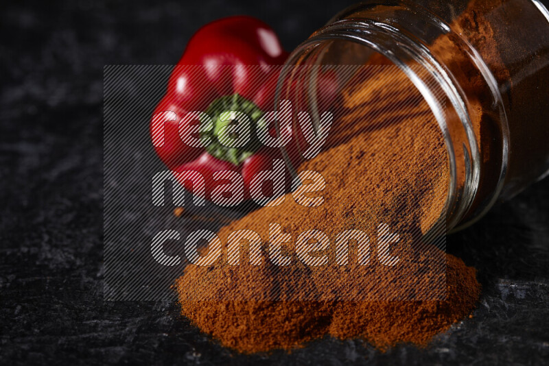 A glass jar full of ground paprika powder flipped with some spilling powder on black background