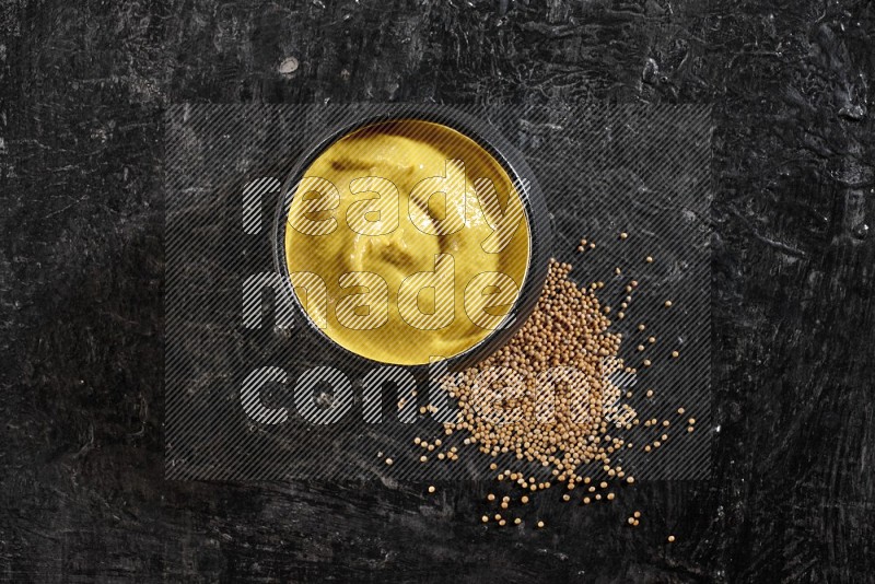 A black pottery bowl full of mustard paste with mustard seeds underneath on textured black flooring