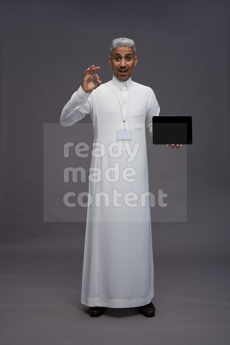 Saudi man wearing thob with neck strap employee badge standing showing tablet to camera on gray background