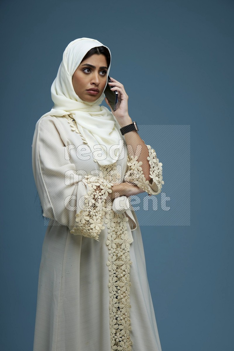 A Saudi woman having a call in a blue background wearing an off-white Abaya Hijab