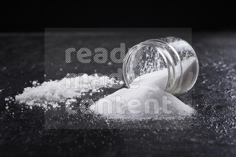 A glass jar full of table salt with some sea salt crystals beside it on a black background