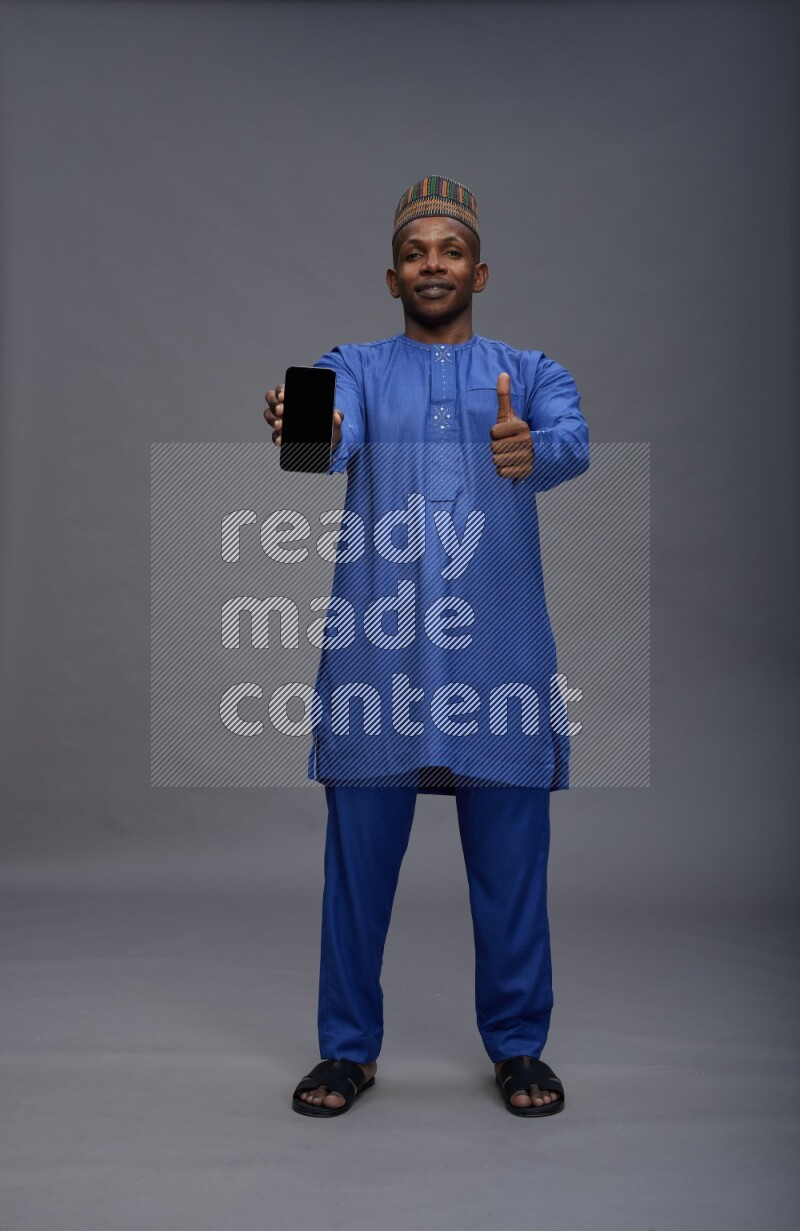 Man wearing Nigerian outfit standing showing phone to camera on gray background