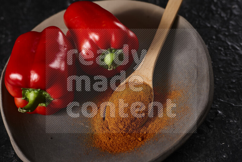 A wooden spoon full of ground paprika powder with red bell peppers, all on a pottery plate on black background