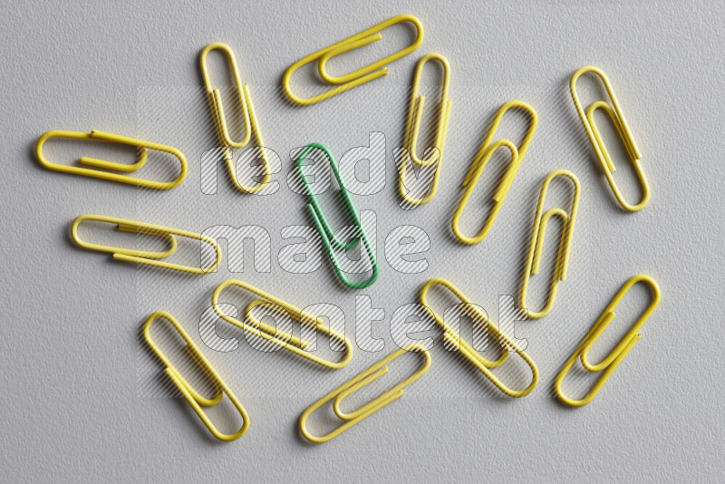 A green paperclip surrounded by bunch of yellow paperclips on grey background