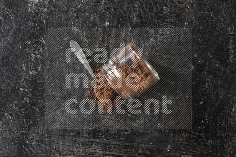 A flipped glass jar and metal spoon full of cloves powder with cloves spread on a textured black flooring