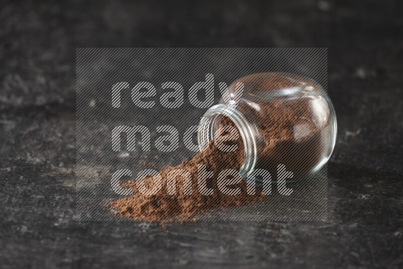 A flipped glass spice jar full of cloves powder and powder came out of it on textured black flooring