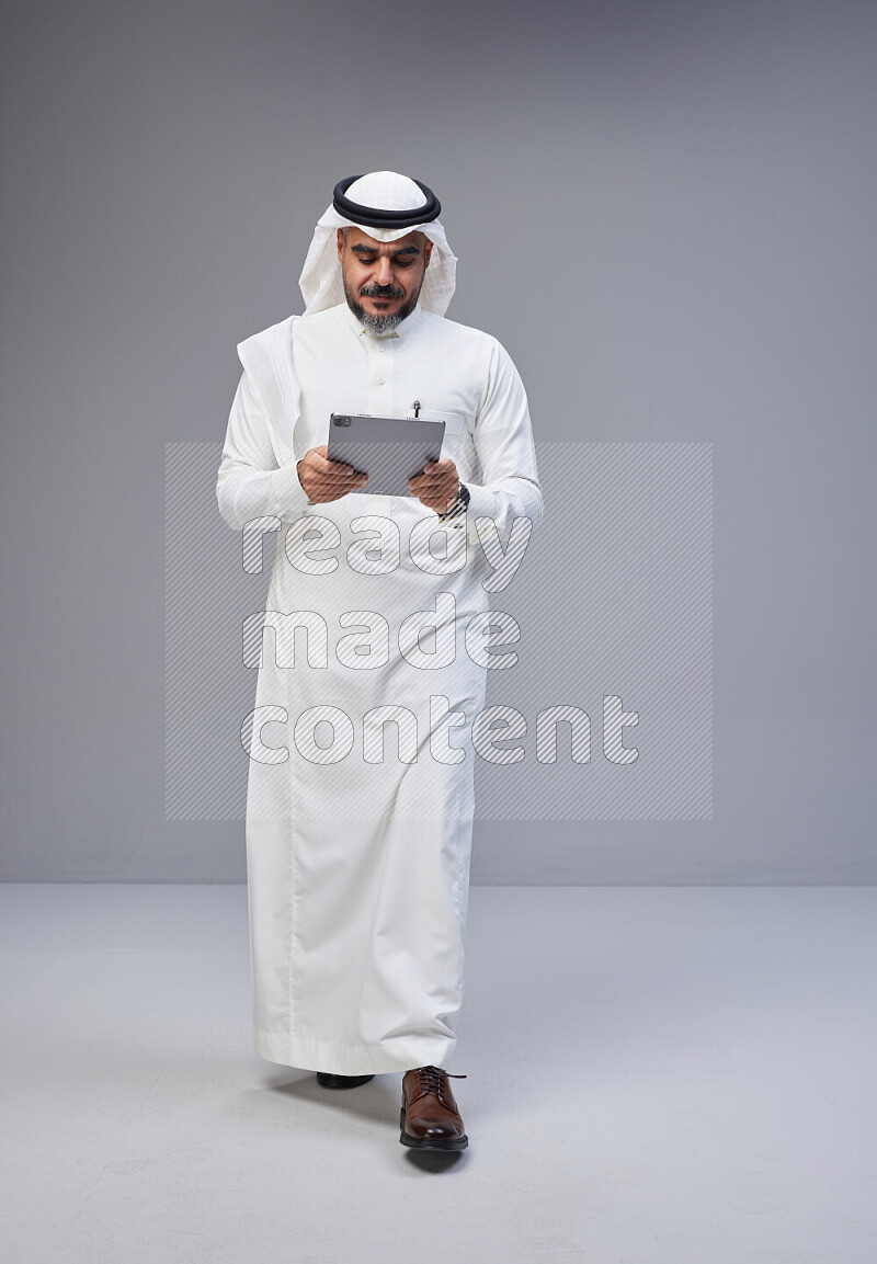 Saudi man Wearing Thob and white Shomag standing working on tablet on Gray background