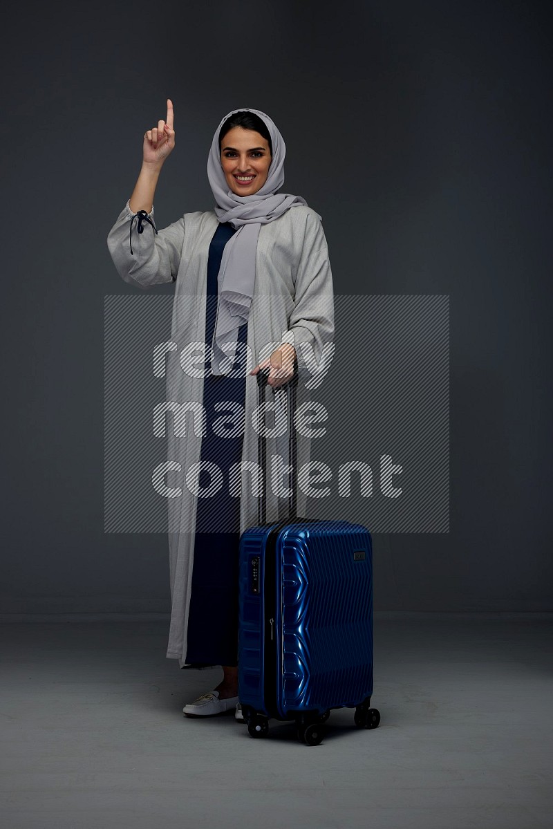 A Saudi woman wearing a light gray Abaya and head scarf standing while holding blue luggage eye level on a grey background