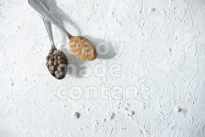 2 metal spoons full of allspice powder and whole balls on a textured white flooring