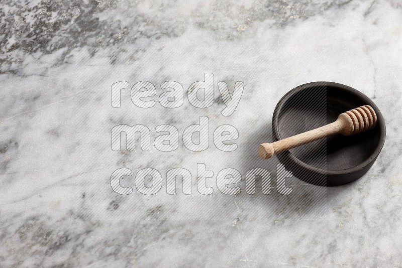 Black Pottery Bowl with wooden honey handle in it, on grey marble flooring, 65 degree angle