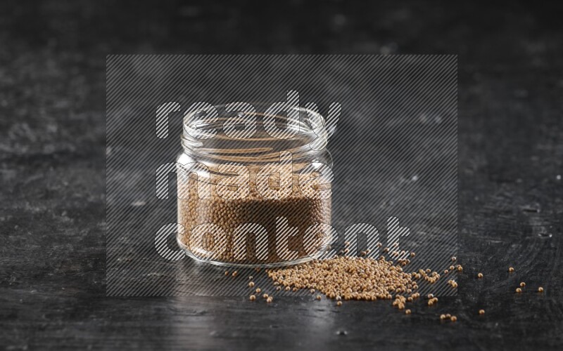A glass jar full of mustard seeds and jar is flipped and seeds spread out on a textured black flooring