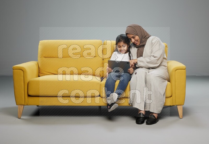 A girl with her mother on yellow sofa and watching on iPad on gray background
