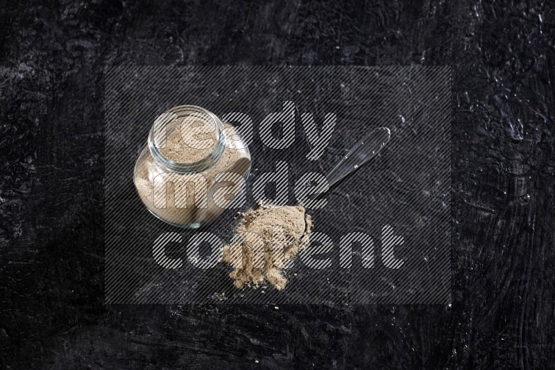 A glass spice jar full of garlic powder with metal spoon on a textured black flooring in different angles