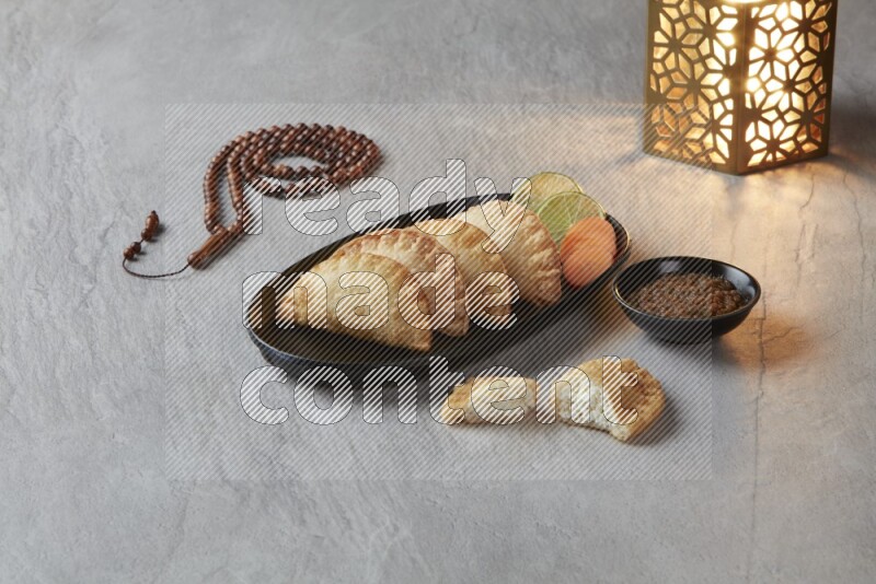 Four fried sambosas in an oval shaped black plate, beside a cut cheese sambosa, a brown misbaha and a golden lantern on a gray background