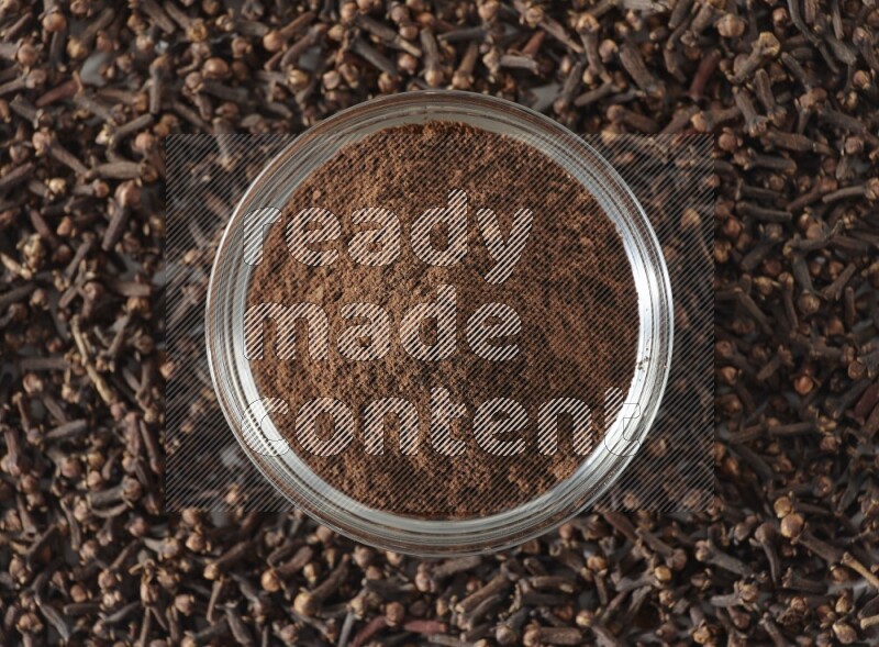 وعاء زجاجي ممتلئ ببودرة القرنفل وحبوب القرنفل متناثرة على أرضية بيضاء