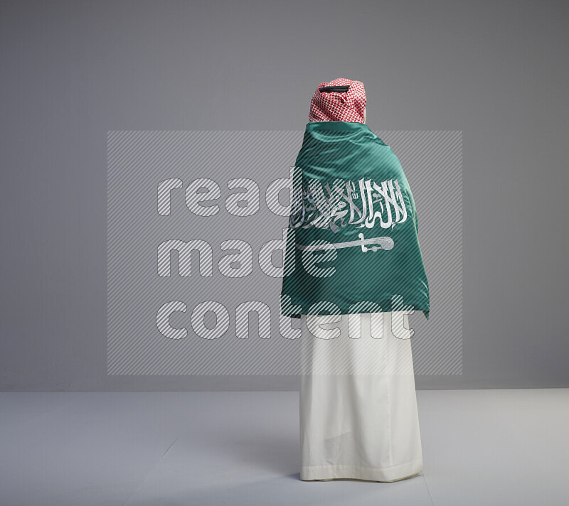A Saudi man standing wearing thob and red shomag wrapping big saudi flag on gray background