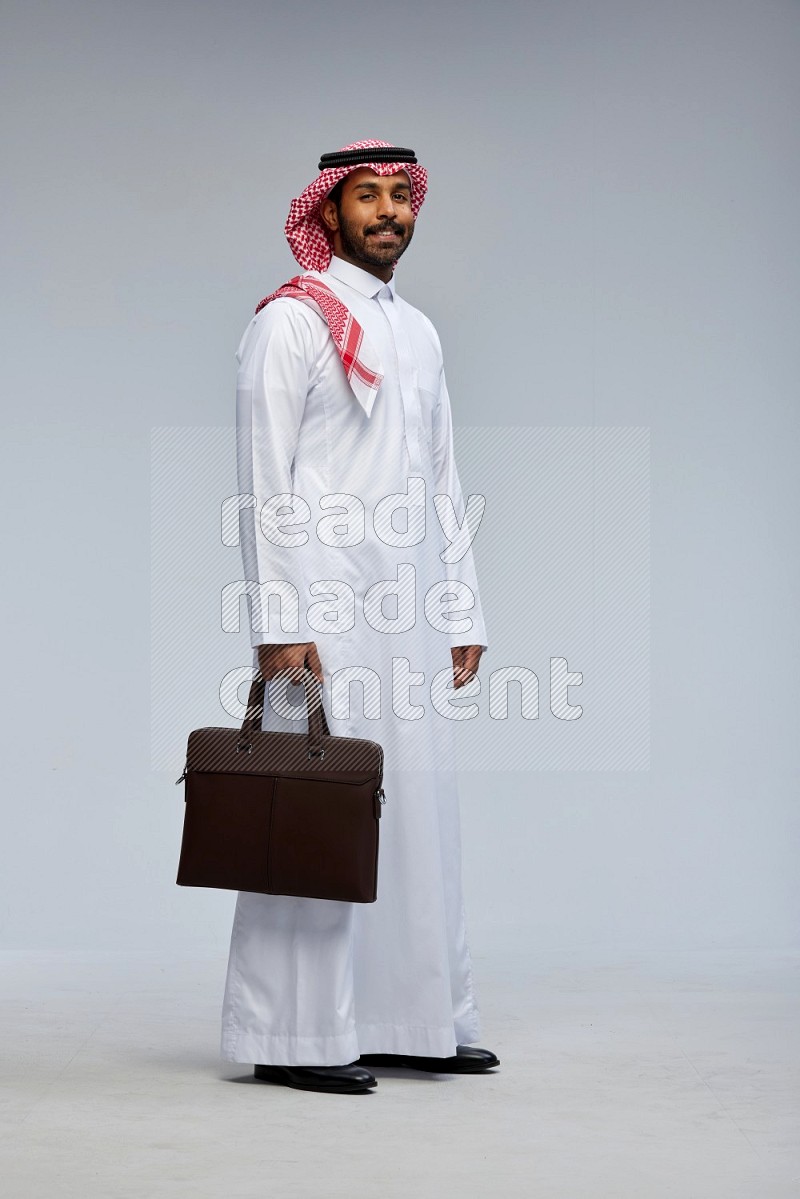 Saudi man Wearing Thob and shomag standing holding bag on Gray background